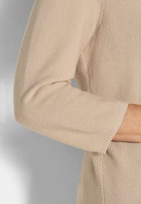 Beiger Strickpullover mit einem gerippten Ärmelbund und einem strukturierten Körper. Nahtdetails entlang des Arms sichtbar. Weiches, leichtes Material.