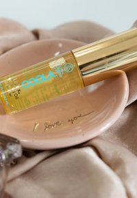 Coola Lipix Hydrating Oil in einer klaren Tube mit einer goldenen Kappe, platziert auf einem rosa Teller, der mit "Ich liebe dich" geprägt ist, vor einem glatten Stoffhintergrund.