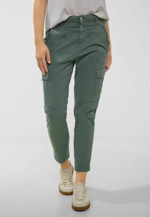 Persoon die groene cargobroek met zijzakken en beige sneakers draagt, staand voor een effen lichte achtergrond.