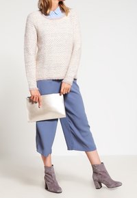 Mujer vistiendo un suéter texturizado rosa, pantalones recortados azules, botas de ante gris hasta el tobillo, sosteniendo un bolso de mano plateado.
