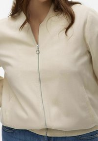 Beige zippered bomberjacka i mjukt material, med rund halsringning, framfickor och en slät silverfärgad dragkedjedetalj.