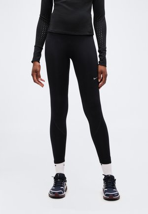 Személy fekete hosszú ujjú felsőt és fekete Nike leggingset visel fehér logóval, fehér zoknival és fekete sportcipővel egyszínű háttér előtt.