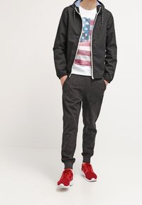 Personne portant un sweat à capuche noir zippé, un t-shirt blanc avec le drapeau américain, un pantalon de jogging à motifs foncés et des chaussures de sport rouges, debout les mains dans les poches.