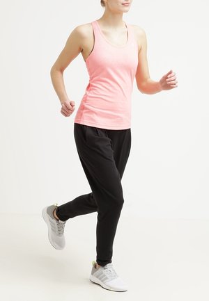 Kobieta w różowym topie bez rękawów, czarnych spodniach dresowych i białych butach sportowych, chodząca lub biegająca na jednolitym jasnym tle.