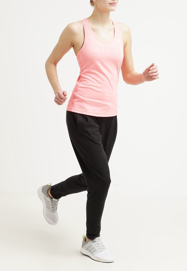 Femme en haut sans manches rose, pantalon de jogging noir et baskets blanches marchant ou faisant du jogging sur un fond clair uni.