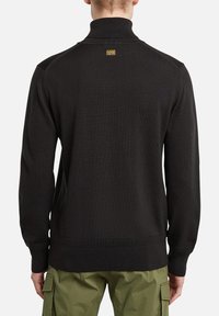 Schwarzer Pullover mit hohem Kragen, gerippter Struktur und Logo-Detail auf der Rückseite; kombiniert mit olivgrünen Cargohosen.