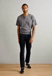 Gray short-sleeve t-shirt, black textured jeans, and black sneakers with white accents. Model stands on a light wooden floor against a plain background.