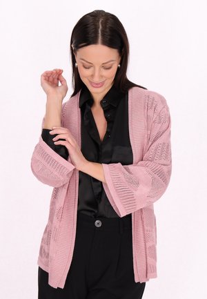 Frau mit dunklem Haar, die Perlenohrringe, eine schwarze Bluse und eine pinke offene Strickjacke trägt, richtet ihren Ärmel während sie nach unten schaut und lächelt.