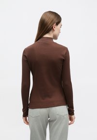 Pruun pikkade varrukatega pluus kõrge kaelusega, valmistatud soonikust kangast. Kannetakse heledate hallide pükstega, vaade tagant.