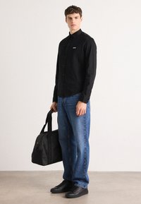 Black button-up shirt with a logo, blue denim jeans, black sneakers, and a black bag with a shoulder strap, set against a neutral background.