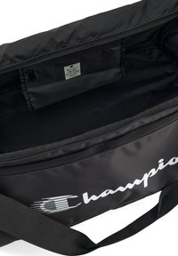 Borsa sportiva nera realizzata in nylon con chiusura a zip. Presenta un logo bianco, una tracolla ampia e una texture liscia. Etichetta interna visibile.