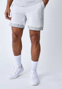 Shorts blancs avec un motif géométrique le long de la taille, arborant un texte de marque gris. Associés à des baskets blanches et des chaussettes.