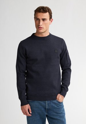 Navy blauwe gebreide trui met een ronde hals, geribbelde manchetten en zoom, en een klein logoplaatje op de borst. Gedragen over een blauwe spijkerbroek.