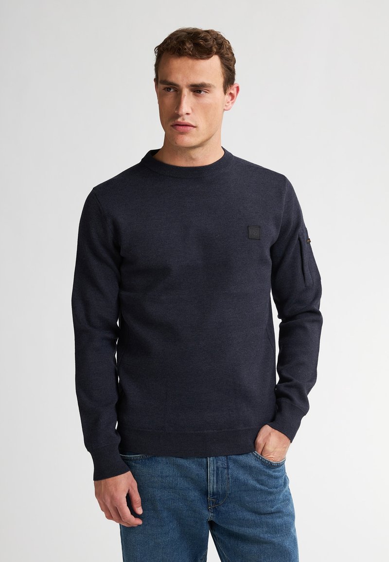 Navy blauwe gebreide trui met een ronde hals, geribbelde manchetten en zoom, en een klein logoplaatje op de borst. Gedragen over een blauwe spijkerbroek.