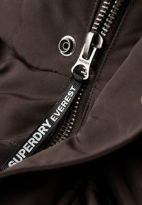Interior de chaqueta de tela marrón con un zipper metálico plateado, que presenta una etiqueta negra con texto blanco que dice "SUPERDRY EVEREST" unida al tirador.