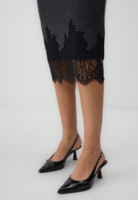 Graues Kleid mit aufwendiger schwarzer Spitzenborte am Saum. Schwarze patentierte Slingback-Schuhe mit spitzem Zehenbereich und schmalem Absatz.