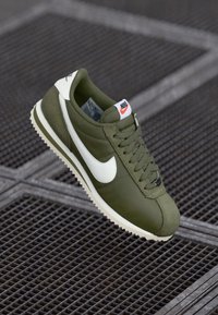 Olive green sneaker with white accents, textured upper, smooth tongue, and classic Nike logo. Features a contrasting cream sole and lace-up design.