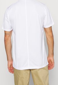 Bílá sportovní košile s krátkým rukávem, hladká textura, má střední šev a malé boční rozparky na lemu.