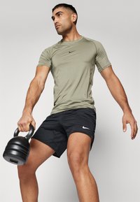 Oliven grønn treningsskjorte med korte ermer, sammen med svarte shorts med en hvit Nike-logo. Modellen holder en svart kettlebell.