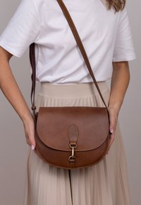 Bolso de hombro de cuero marrón con cierre de solapa, que presenta un clasp metálico y una pestaña decorativa de cuero, sobre un fondo claro.