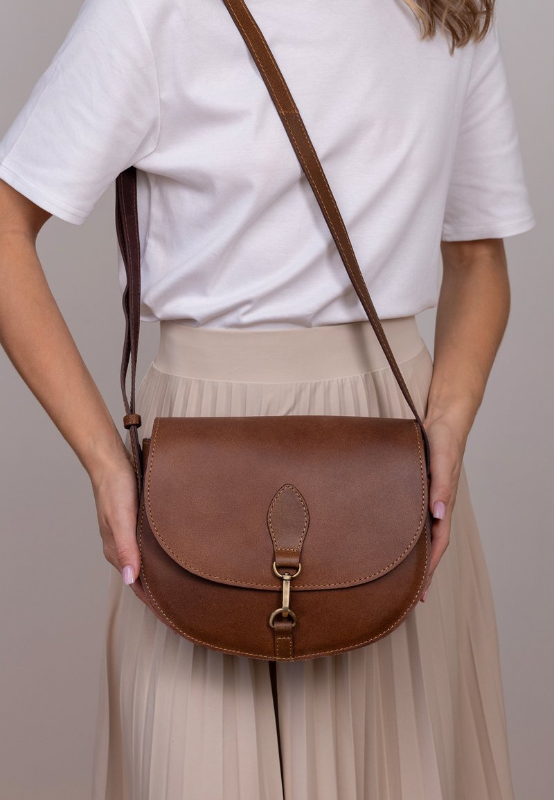 Brun læder crossbody taske med klaplukning, der har en metalklap og en dekorativ læderflap, anbragt på en lys baggrund.