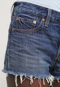 Los pantalones cortos de mezclilla presentan una tela azul desgastada, dobladillo deshilachado, diseño de cinco bolsillos, remaches de bronce y detalles de costura visibles.