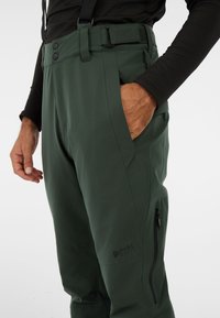 Groene waterdichte broek met verstelbare tailleband, zijzakken en ritsen. Heeft een logo op het been en een gestructureerde afwerking.