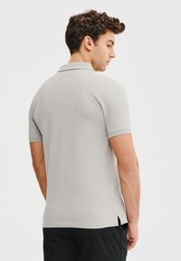 Jasnoszara koszulka polo z krótkimi rękawami, o teksturowanej tkaninie i klasycznym kołnierzyku, posiada boczne rozcięcia u dołu dla swobody ruchów.