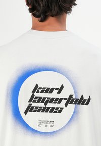 Hvid t-shirt med korte ærmer, der har et cirkulært blåt gradientdesign og fed sort tekst: "karl lagerfeld jeans" og "NEW ECLIPSE."
