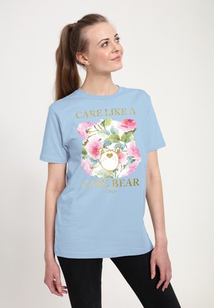 T-shirt in cotone azzurro chiaro con un grafico di un Care Bear circondato da rose rosa e testo dorato che recita "Prenditi Cura Come un Care Bear."