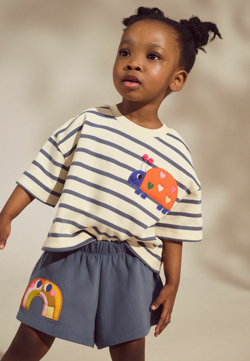 Junges Kind mit geflochtenem Haar trägt ein gestreiftes Shirt mit einem Marienkäfer-Patch und graue Shorts mit einem Regenbogen-Patch, steht vor einem einfachen Hintergrund.