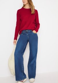Czerwony sweter w prążki, szerokie ciemnoniebieskie dżinsy i jasne sneakersy. Modelka trzyma torebkę na ramię w kolorze kremowym.