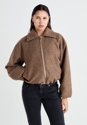 Veste en shearling marron, duveteuse, avec un col, une fermeture éclair devant et des manches volumineuses, associée à un jeans noir ajusté.
