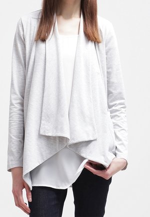 Mujer que lleva un cárdigan gris claro abierto por delante sobre una blusa blanca combinado con pantalones oscuros, de pie frente a un fondo liso.