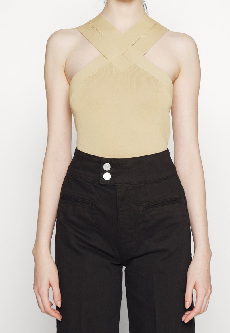 Femme portant un haut beige sans manches avec des bretelles croisées et un pantalon noir taille haute avec deux boutons à l’avant.