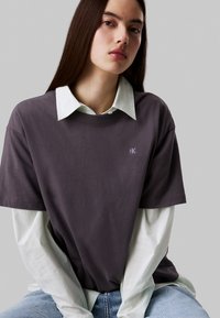 Camiseta de algodón gris superpuesta a una camisa blanca de rayas con cuello, que presenta un pequeño logotipo azul en el pecho y jeans de mezclilla.