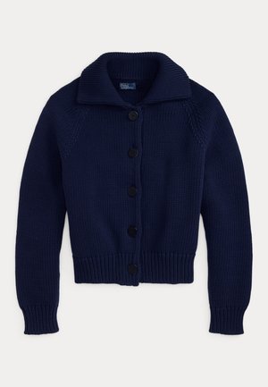 Femeie purtând un cardigan bleumarin cu nasturi peste o cămașă albă, pantaloni largi bej, ținând o geantă maro, stând desculță pe fundal neutru.