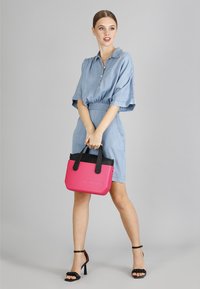 Borsa a tracolla rosa e nera con superficie texturizzata, manici corti e chiusura con zip, tenuta da un modello in un vestito di jeans su uno sfondo neutro.
