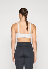 Svetlo siva športna modrček z nastavljivimi naramnicami, gladka tkanina, z zapiranjem na pasu, v kombinaciji z visokimi črnimi leggingami z detajli šivanja.