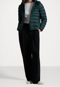 Doudoune vert foncé avec un matelassage horizontal, fermeture éclair et capuche, associée à un pantalon noir ample et des baskets blanches.