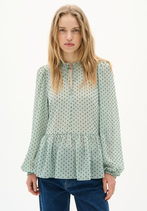 Grön blus med prickar, peplumslut, lätt genomskinlig textur, långa puffärmar och en nyckelhålsringning med en knytdetalj.