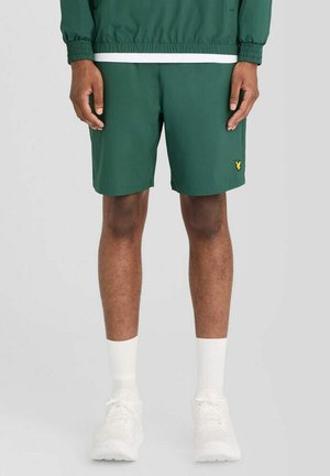 Man draagt groene korte broek met een geel logo, witte sokken, witte sneakers en een bijpassend groen jasje met elastische boorden.