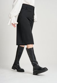 Stivali neri al ginocchio con suola spessa, realizzati in pelle liscia. Il design presenta una cucitura verticale e un tassello per la mano nella parte superiore.