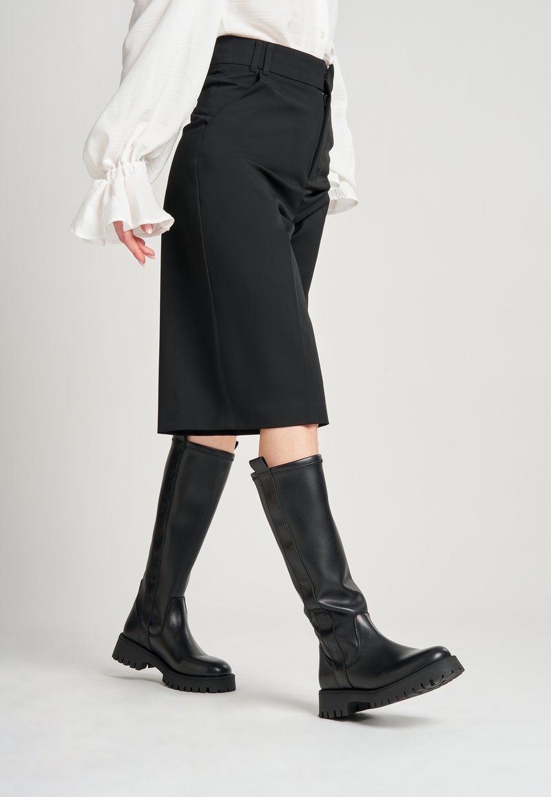 Stivali neri al ginocchio con suola spessa, realizzati in pelle liscia. Il design presenta una cucitura verticale e un tassello per la mano nella parte superiore.