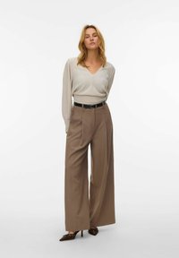 Lichtbeige geplooide blouse met een V-hals, gecombineerd met brede tan pantalons en een zwarte riem. Bruine heeled mules met studaccenten maken de look af.
