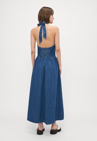 Denim maxi kjole med halterneck-design, åpen rygg, samlet midje og en flytende skjørt. Blå farge med en glatt tekstur.