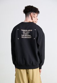 Černá mikina se potištěným textem na zádech, s logem Timberland a detaily produktu, s žebrovanými manžetami a lemem.