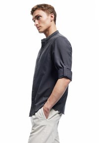 Donkergrijze button-up shirt met een staande kraag en opgerolde mouwen, gedragen door een model met handen in de zakken, gecombineerd met lichtgekleurde broek.