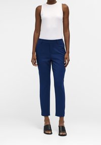 Marineblauwe op maat gemaakte broeken met voorkruiken en een slim fit ontwerp, gecombineerd met een witte mouwloze top en zwarte sandaal-slippers.