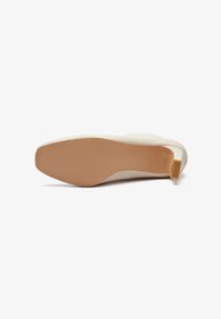 Zapato de tacón beige con textura suave, punta redondeada y un acento amarillo en el tacón. Suela plana de color claro.